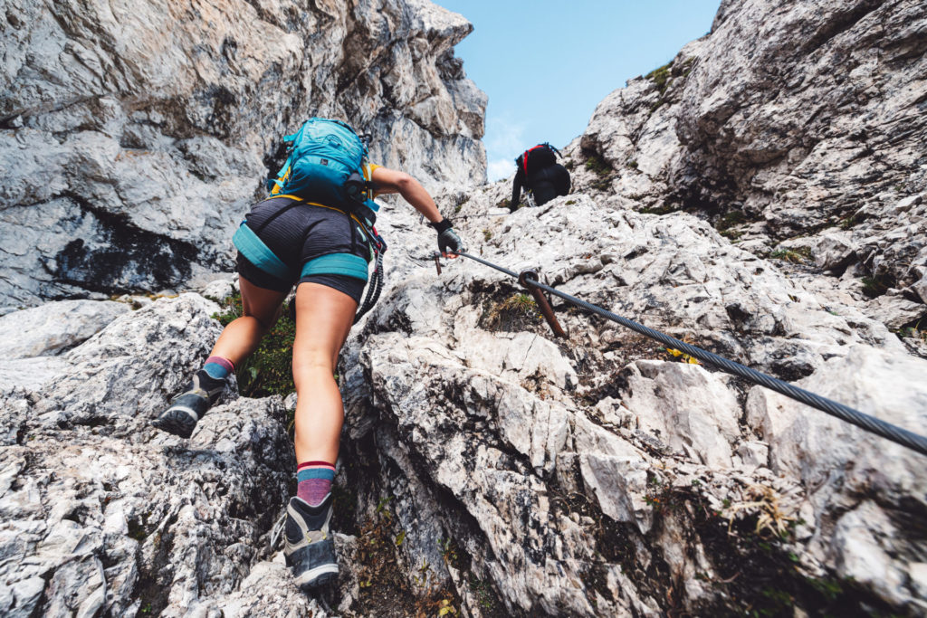 View from the back looking up at woman mountaineer in shorts climbing up on via ferrata trail in the Alps
